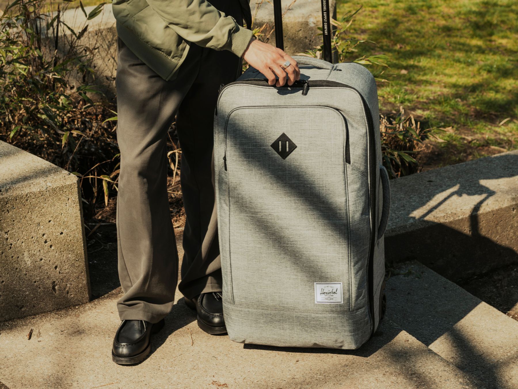 A shot of a person unzipping a Herschel Heritage® Softshell Medium Luggage in Raven Crosshatch in a park.