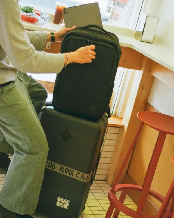 a grey large herschel heritage hardshell luggage with a luggage belt sits in the corner of a cafe. Someone is putting a laptop into a black kaslo daypack that is sitting on top of the luggage.